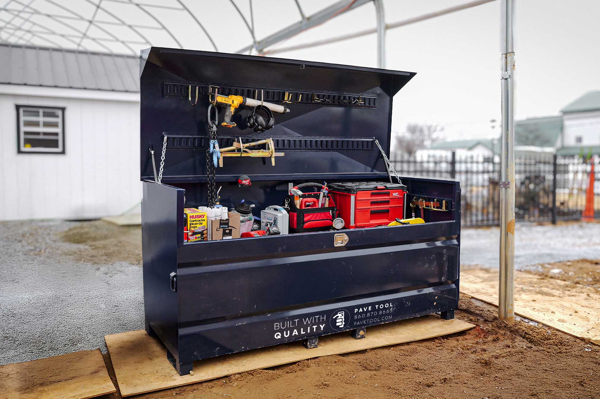 A large, industrial storage container known as Quick-E-BARBoxx, which is black in color, is open and showing various tools and equipment organized inside it. The container is placed outdoors on a wooden palette, and there is a building in the background.